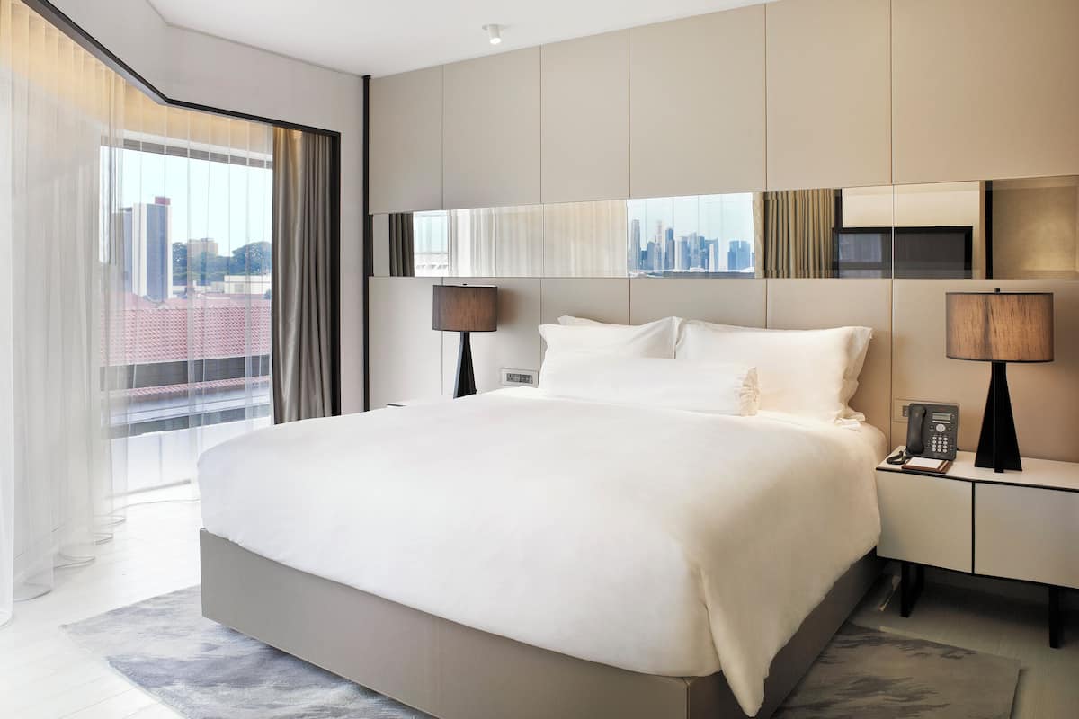 A modern hotel room with a large bed, a city view, a desk with a phone, and a lamp.