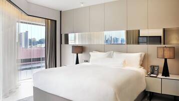 A modern hotel room with a large bed, a city view, a desk with a phone, and a lamp.