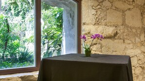 Una mesa con mantel oscuro, un florero con flores moradas y vistas a la vegetación a través de una ventana con un muro de piedra.