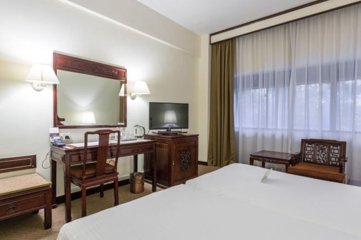 A hotel room with a bed, a desk with a chair, a television, a mirror, and a window with curtains.