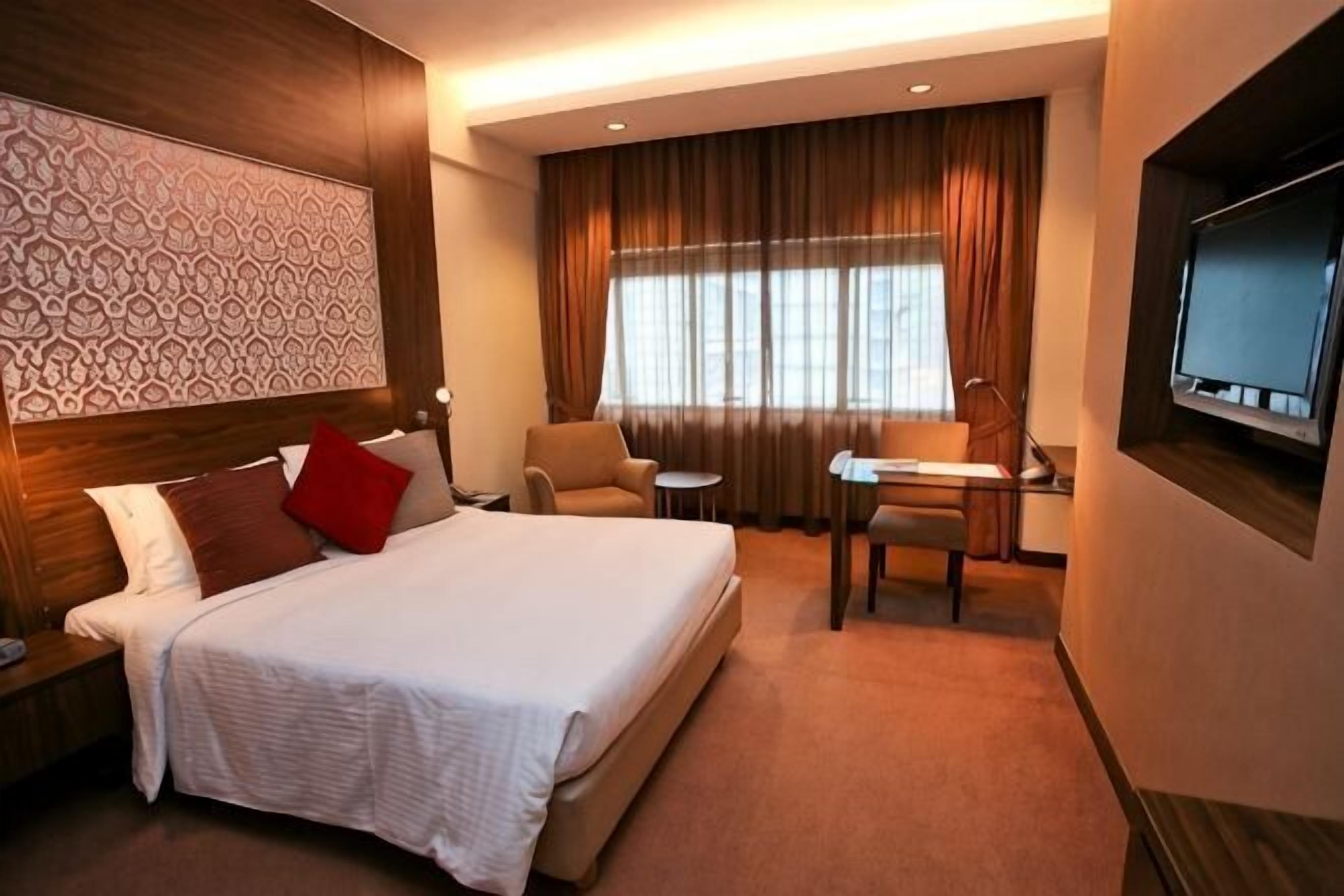Ein Hotelzimmer mit einem großen Bett, einem Schreibtisch, einem Sessel, einem Fernseher und einem Fenster mit Vorhängen.