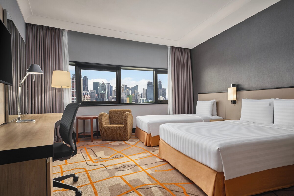 A hotel room with a desk, two beds, a chair, a small table, and a view of the city skyline through large windows.