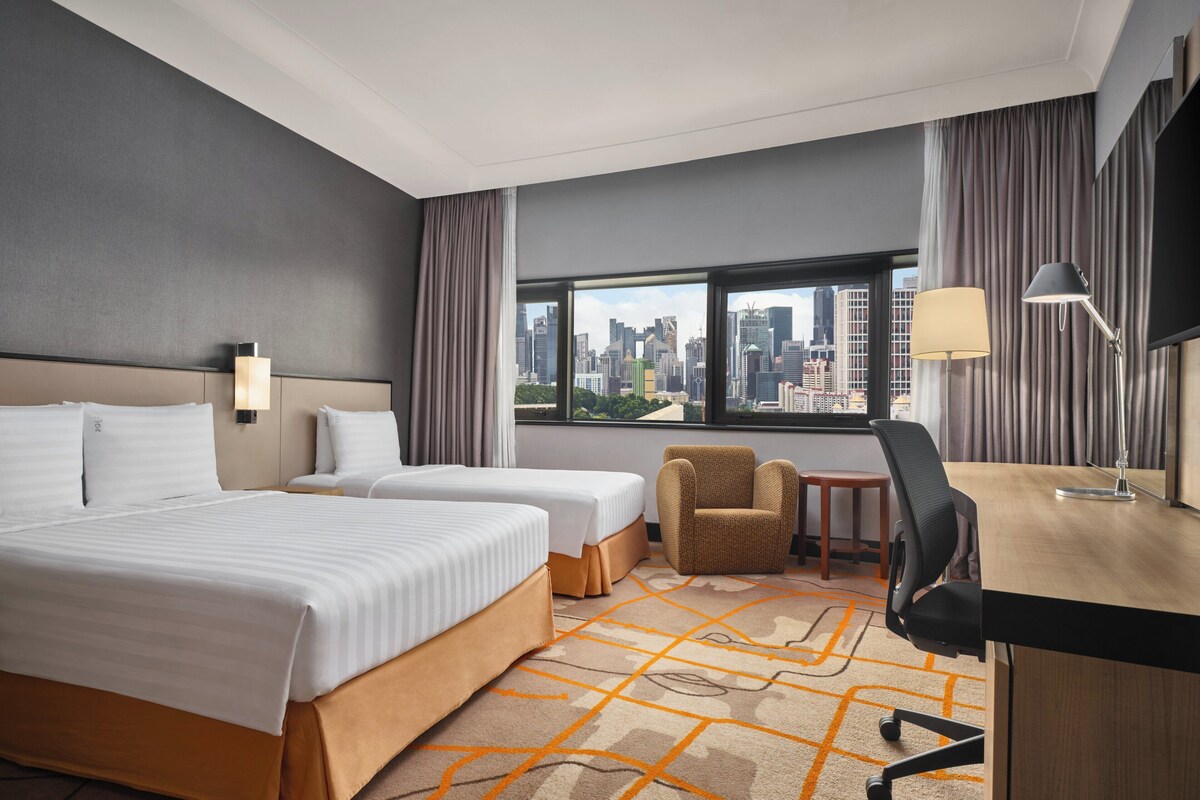 A hotel room with two beds, a desk, a chair, a TV, and a large window with a city view.