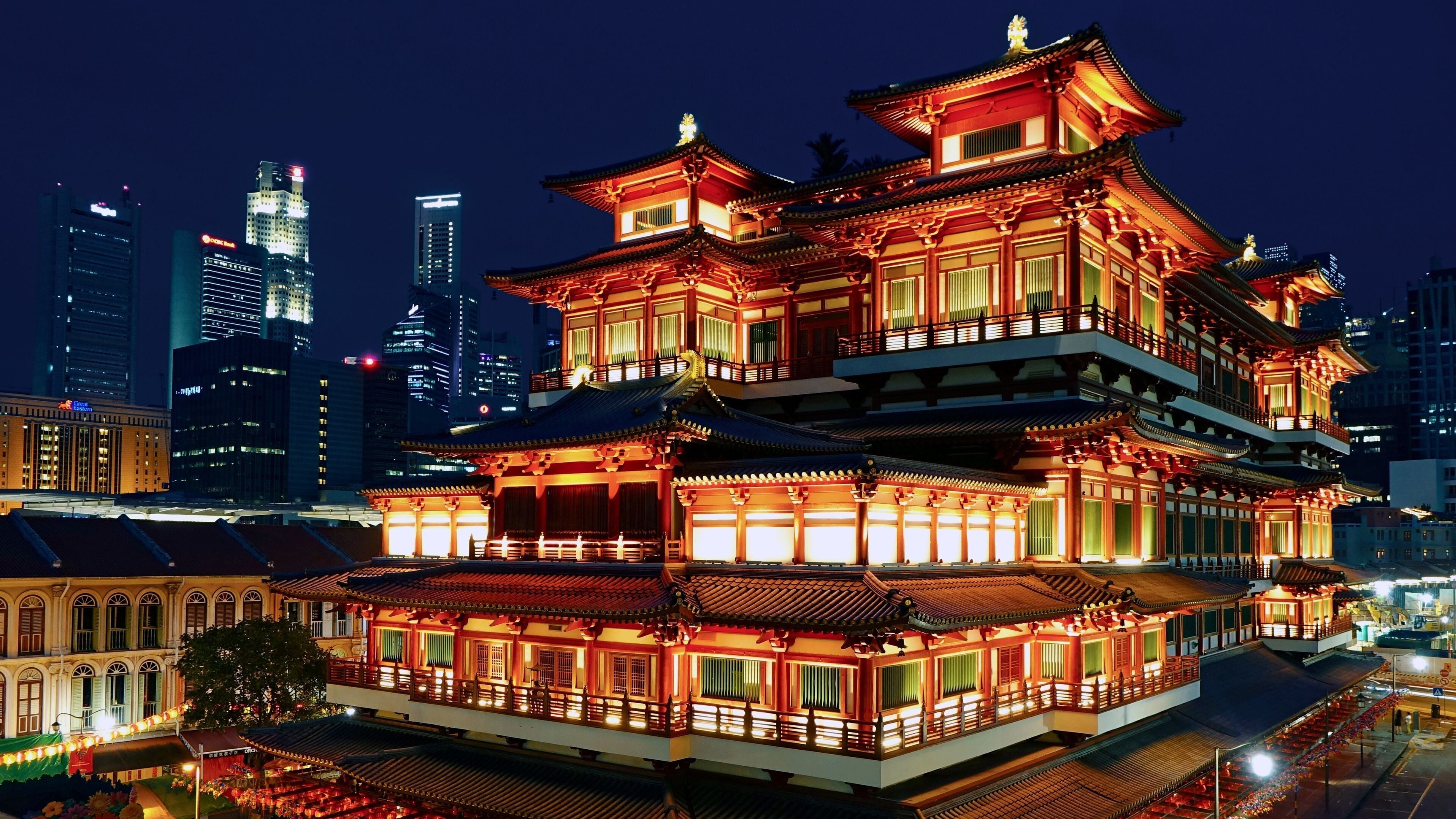 Un bâtiment traditionnel à plusieurs étages, doté de balustrades illuminées, se dressant sur le fond d’un paysage urbain moderne la nuit.