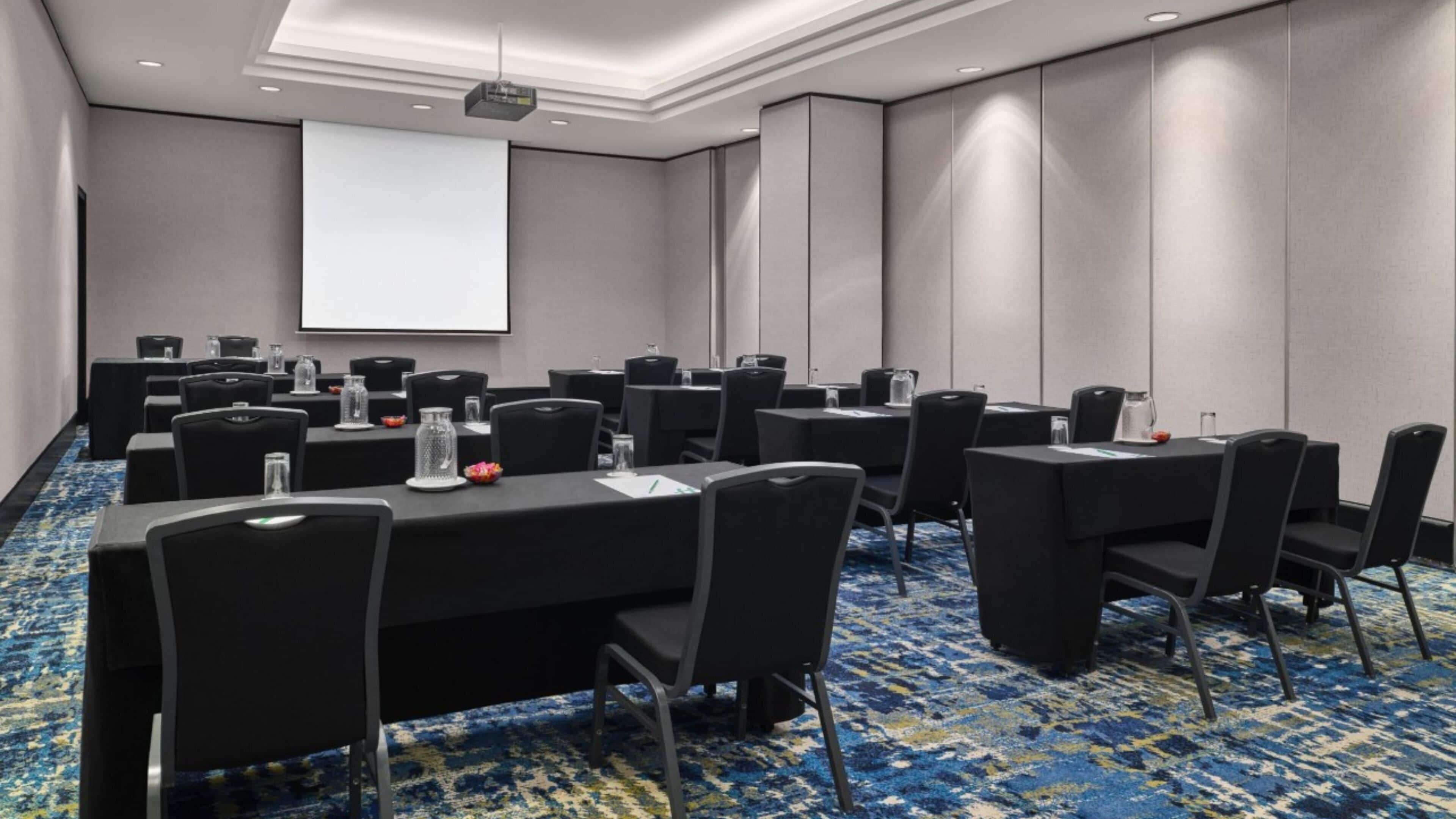 A conference room with multiple tables and chairs, a projector screen, and a carpeted floor.