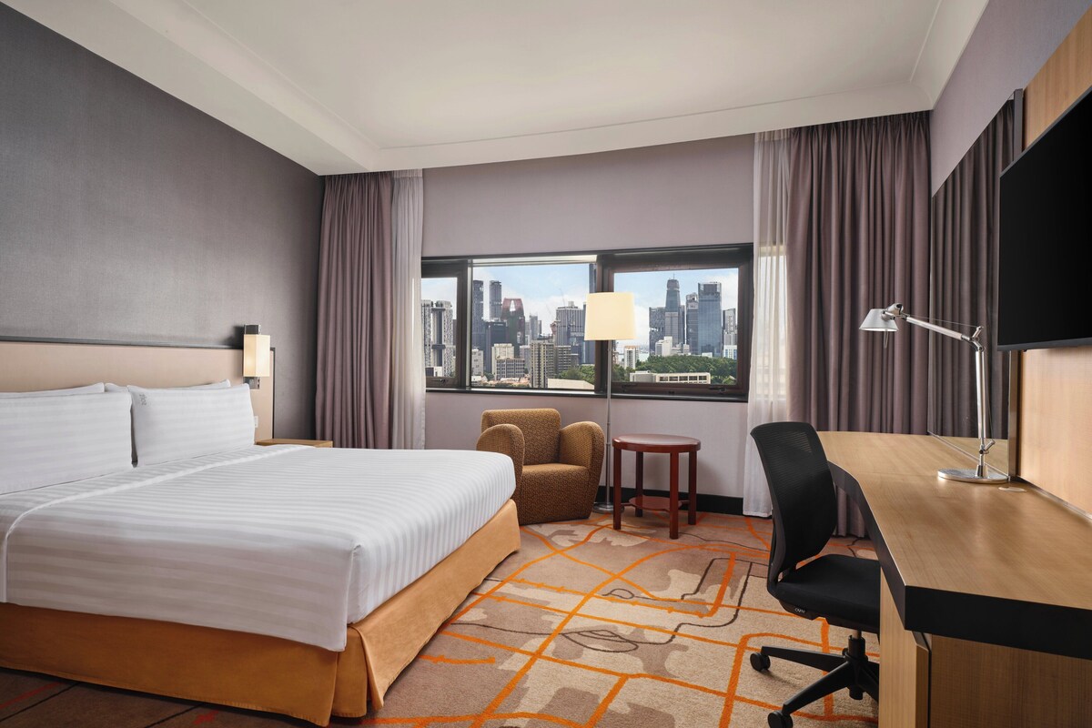 A hotel room with a large bed, a desk, a chair, a small table, a lamp, and a view of a cityscape through a large window.