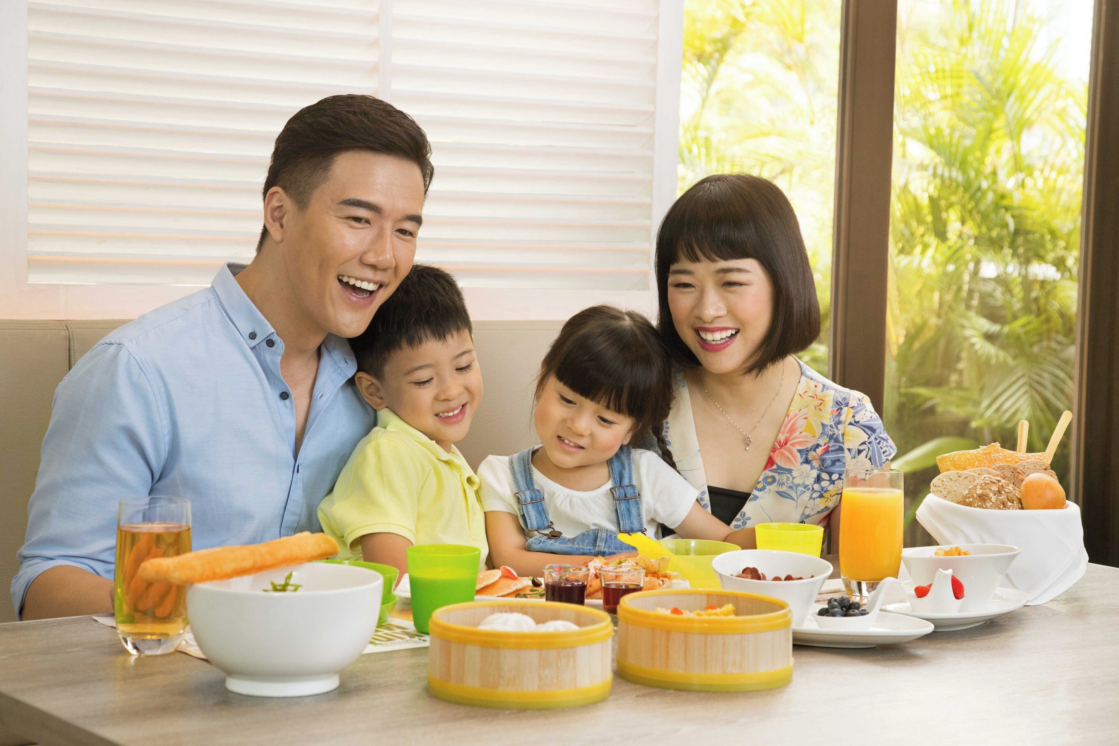 Uma família jantando à mesa, com diversos pratos, bebidas e vista para a janela.