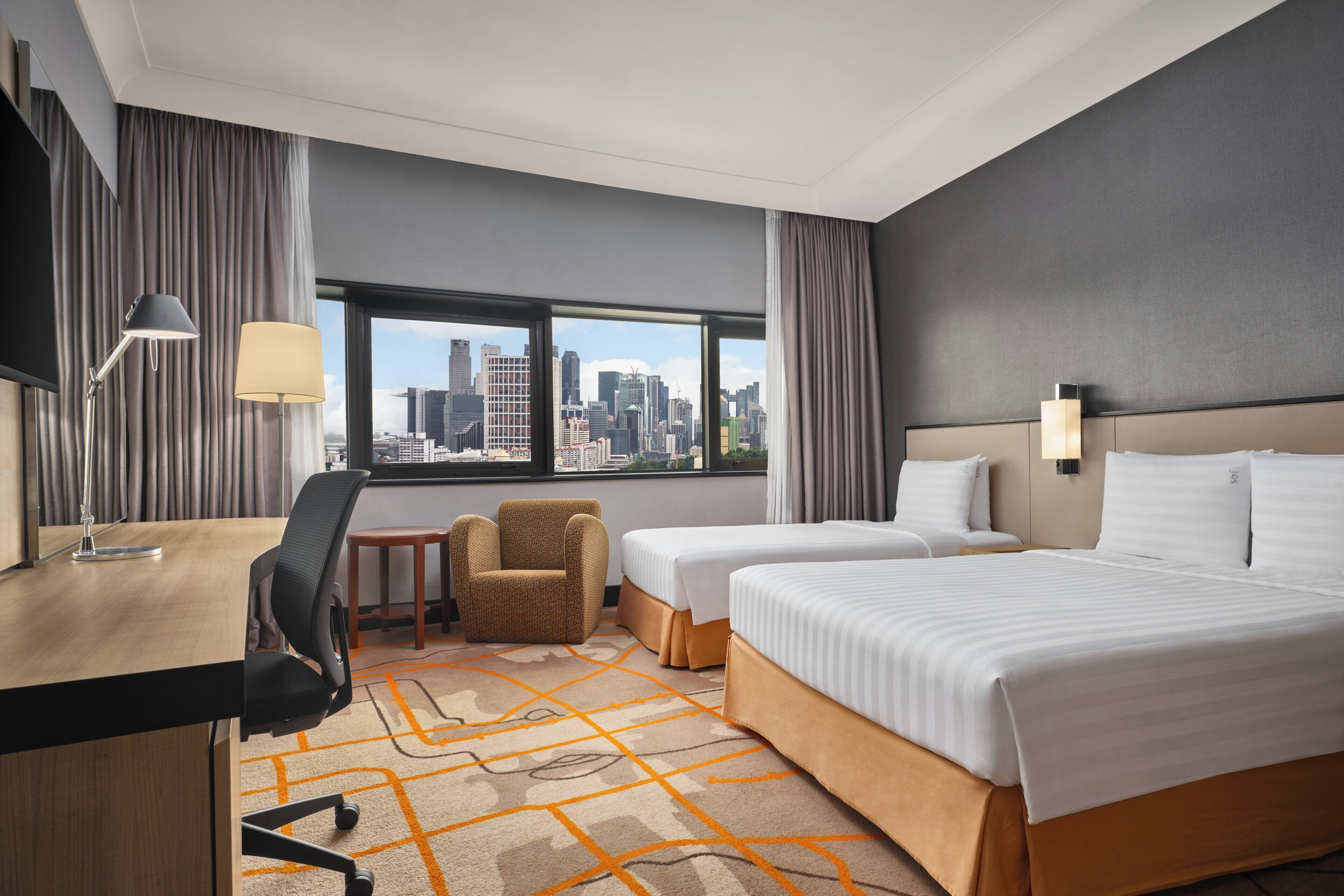 A hotel room with a city view, a desk with a lamp, two beds, a chair, and a small table.