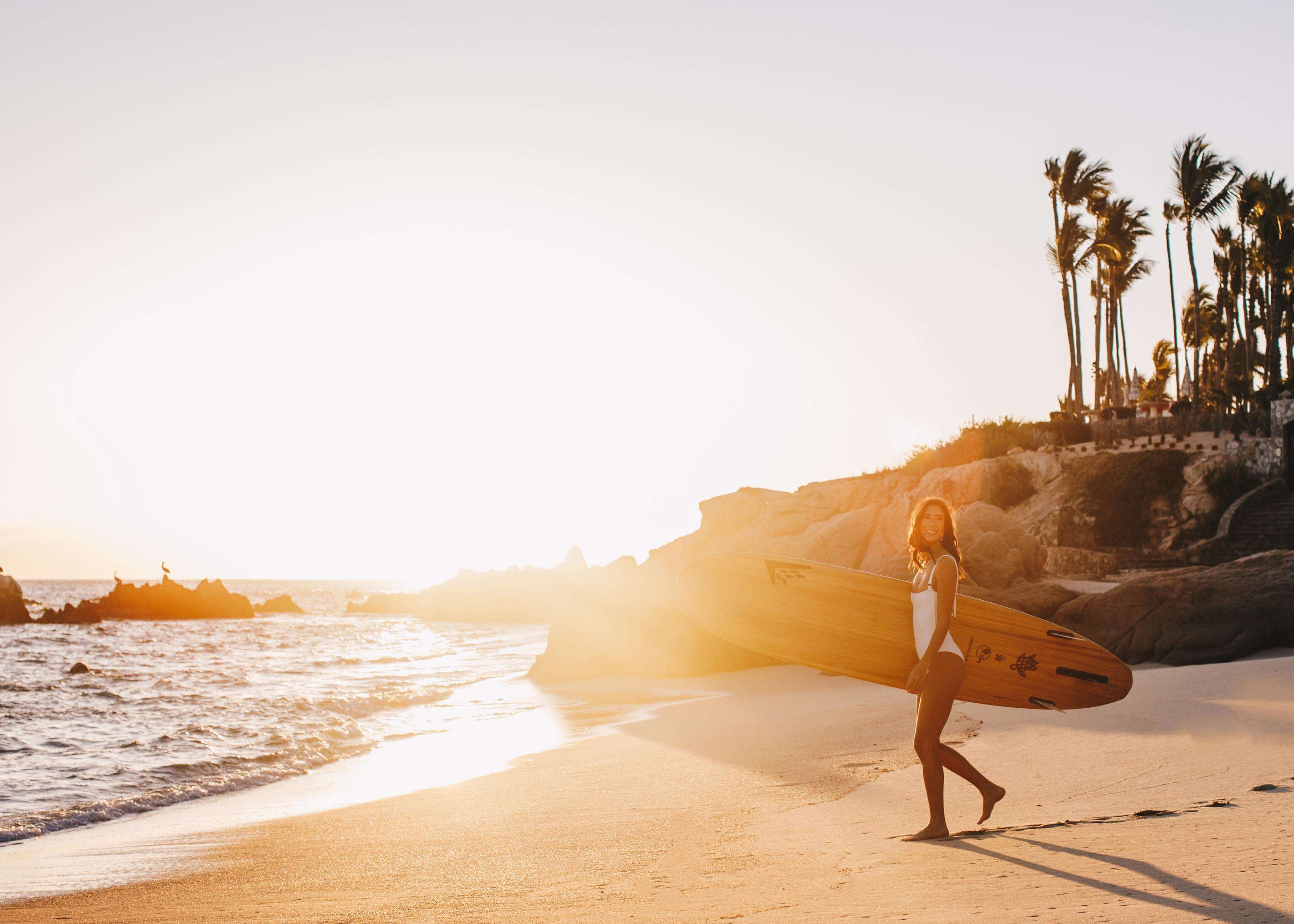 A beach scene with a person carrying a surfboard, palm trees, and a rocky coastline.