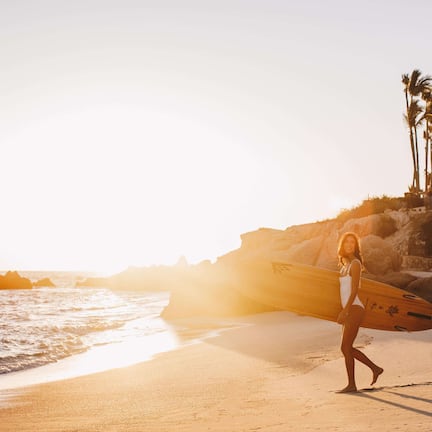 A beach scene with a person carrying a surfboard, palm trees, and a rocky coastline.