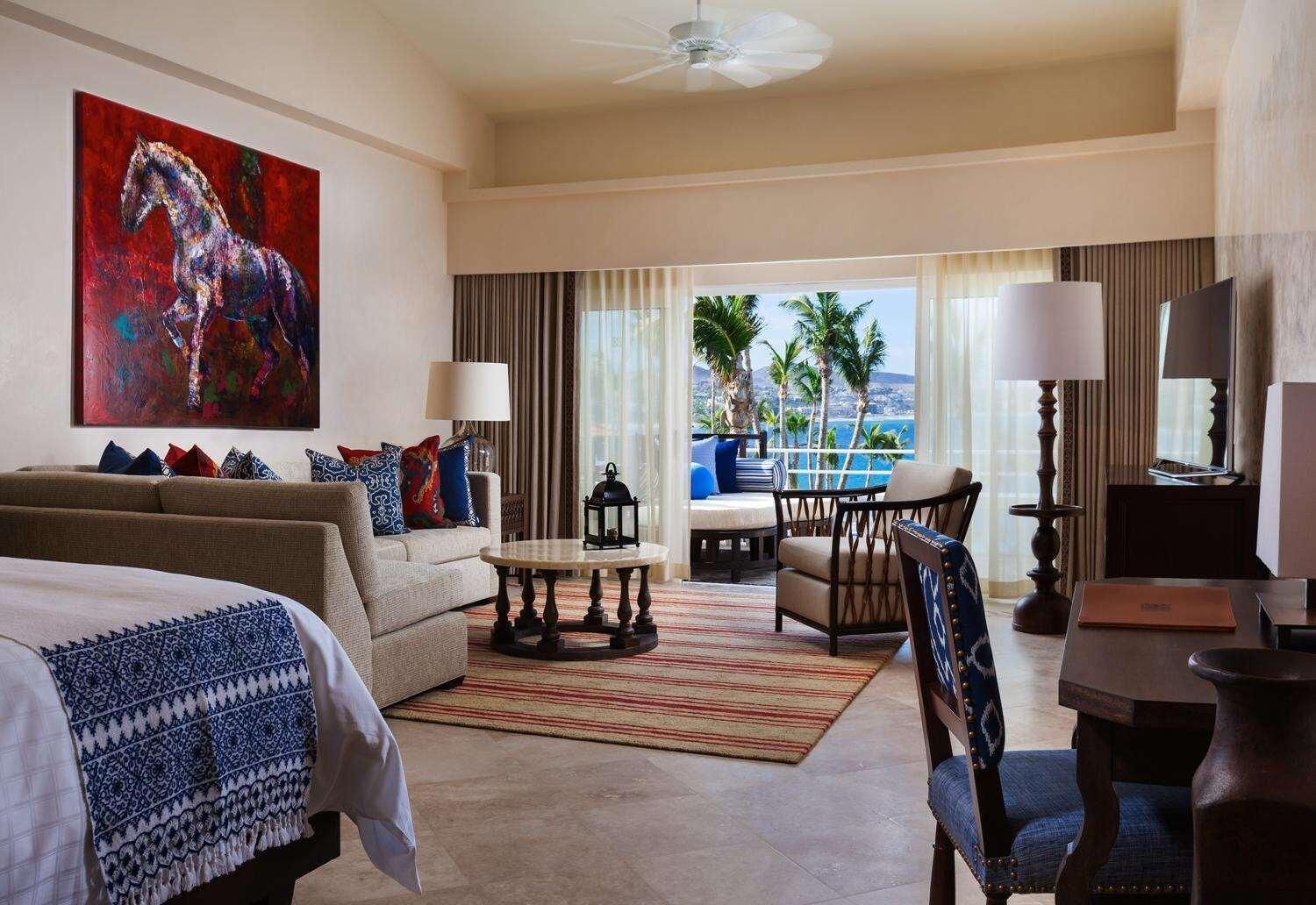 A hotel room with a bed, a sofa, a dining table, a chair, a coffee table, a painting, and a view of the ocean.