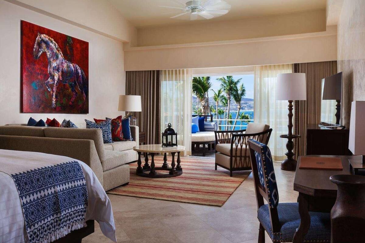 A hotel room with a bed, a sofa, a dining table, a chair, a coffee table, a painting, and a view of the ocean.