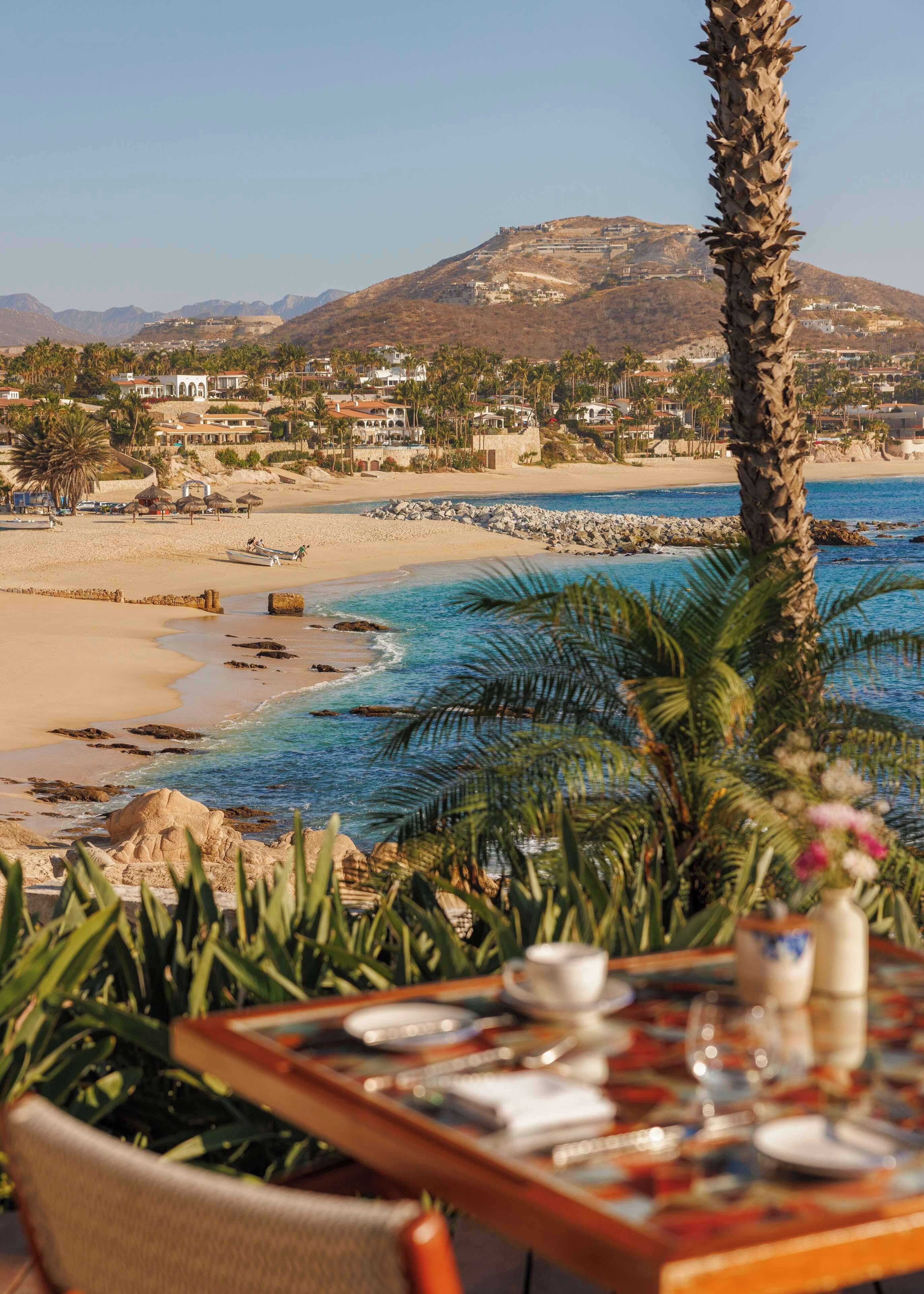 A beachfront dining table with a view of a beach, palm trees, and a mountain in the background.