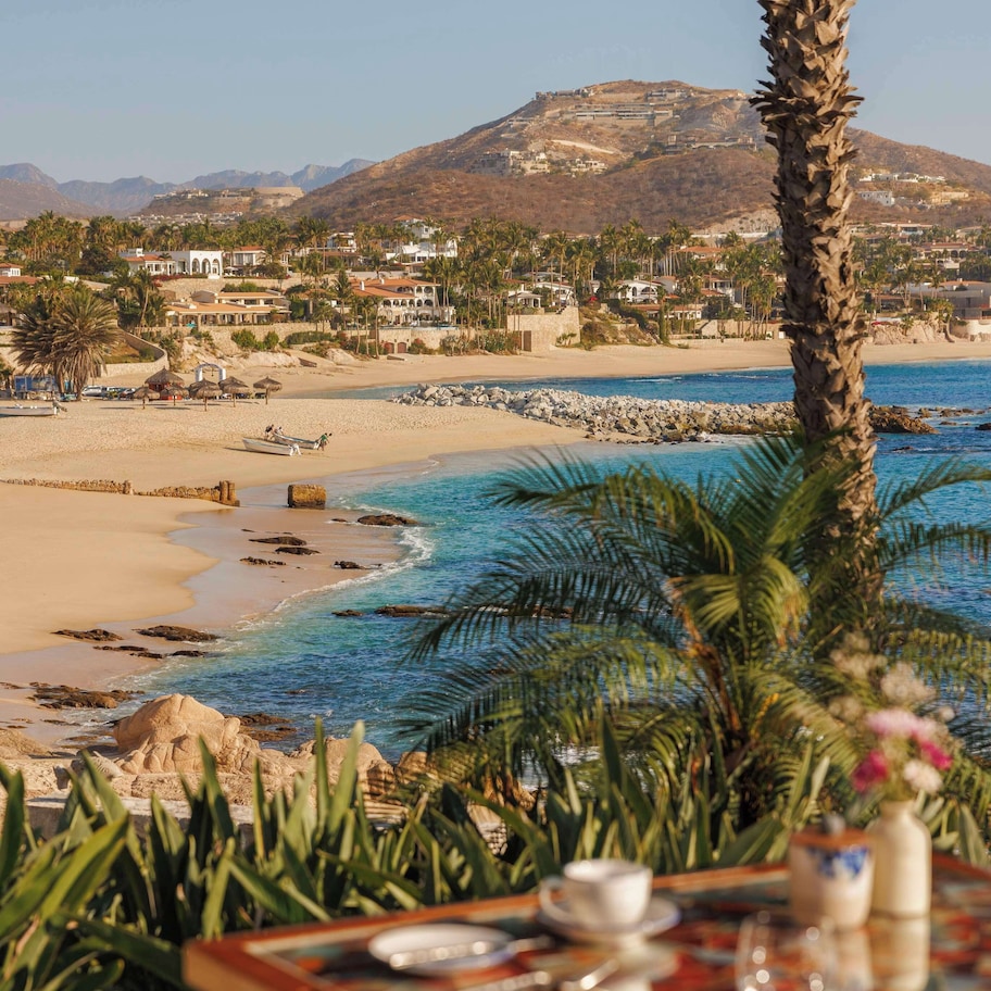 A beachfront dining table with a view of a beach, palm trees, and a mountain in the background.