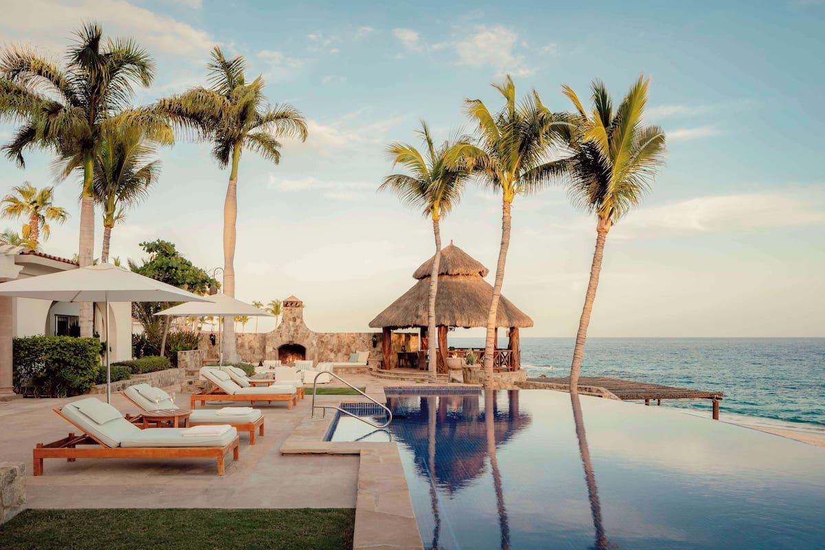 A poolside area with lounge chairs, a thatched-roof structure, and palm trees.