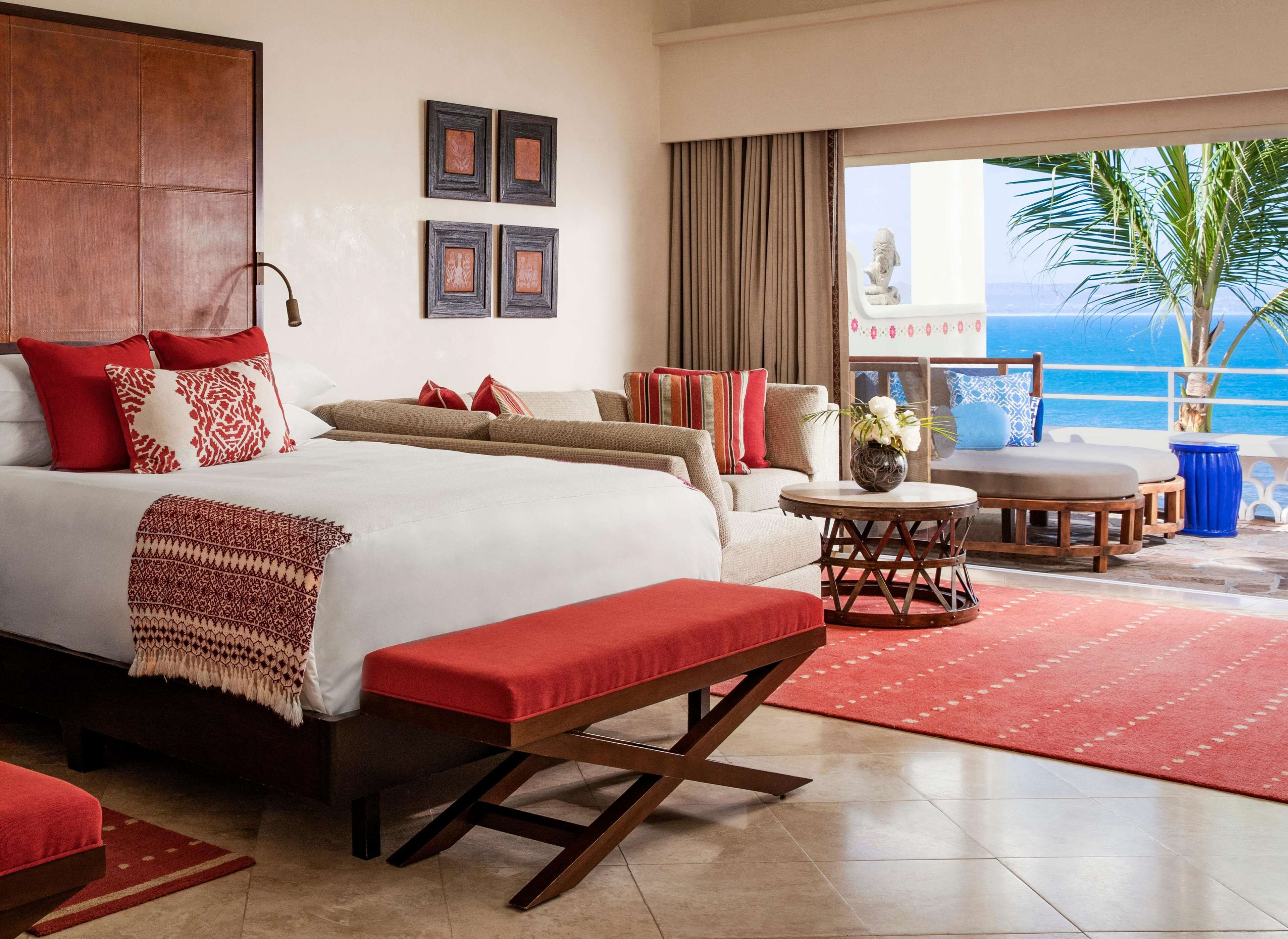 A hotel room with a bed, a bench, a table, and a view of the ocean.