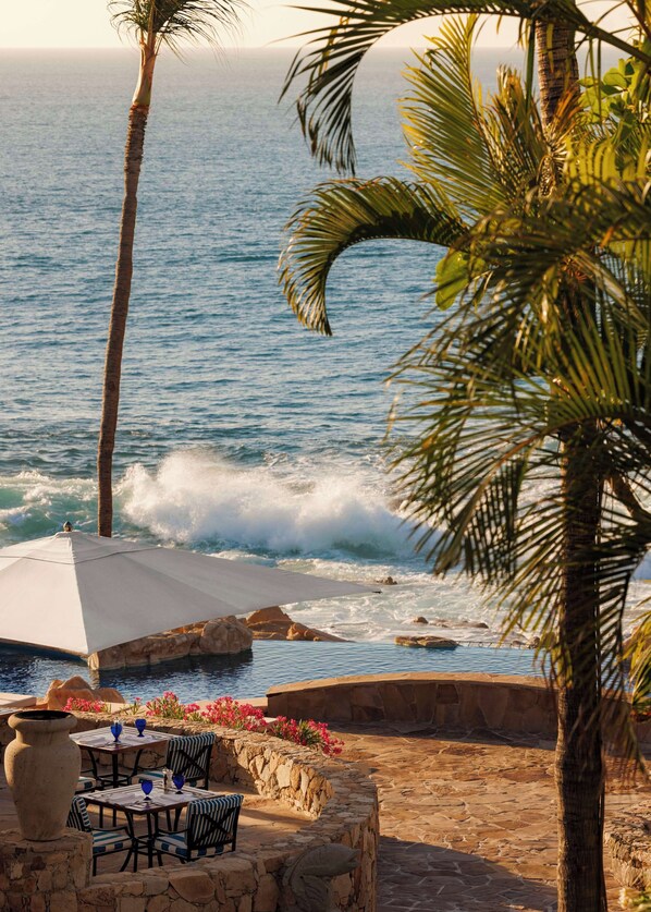 A coastal outdoor seating area with tables and chairs, a stone wall, and a view of the ocean.
