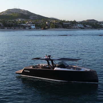 A sleek, modern yacht with a covered deck, anchored near a coastal town with hills in the background.