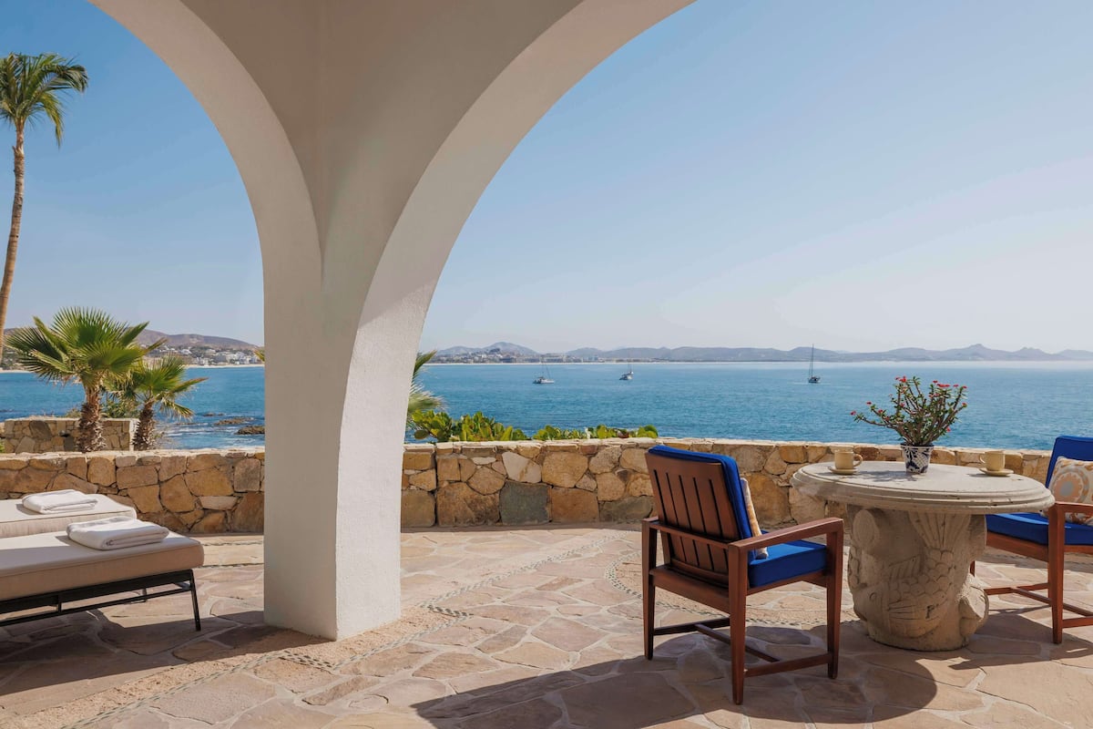 A stone patio with a lounge chair, a small table with a vase, and a view of the sea.