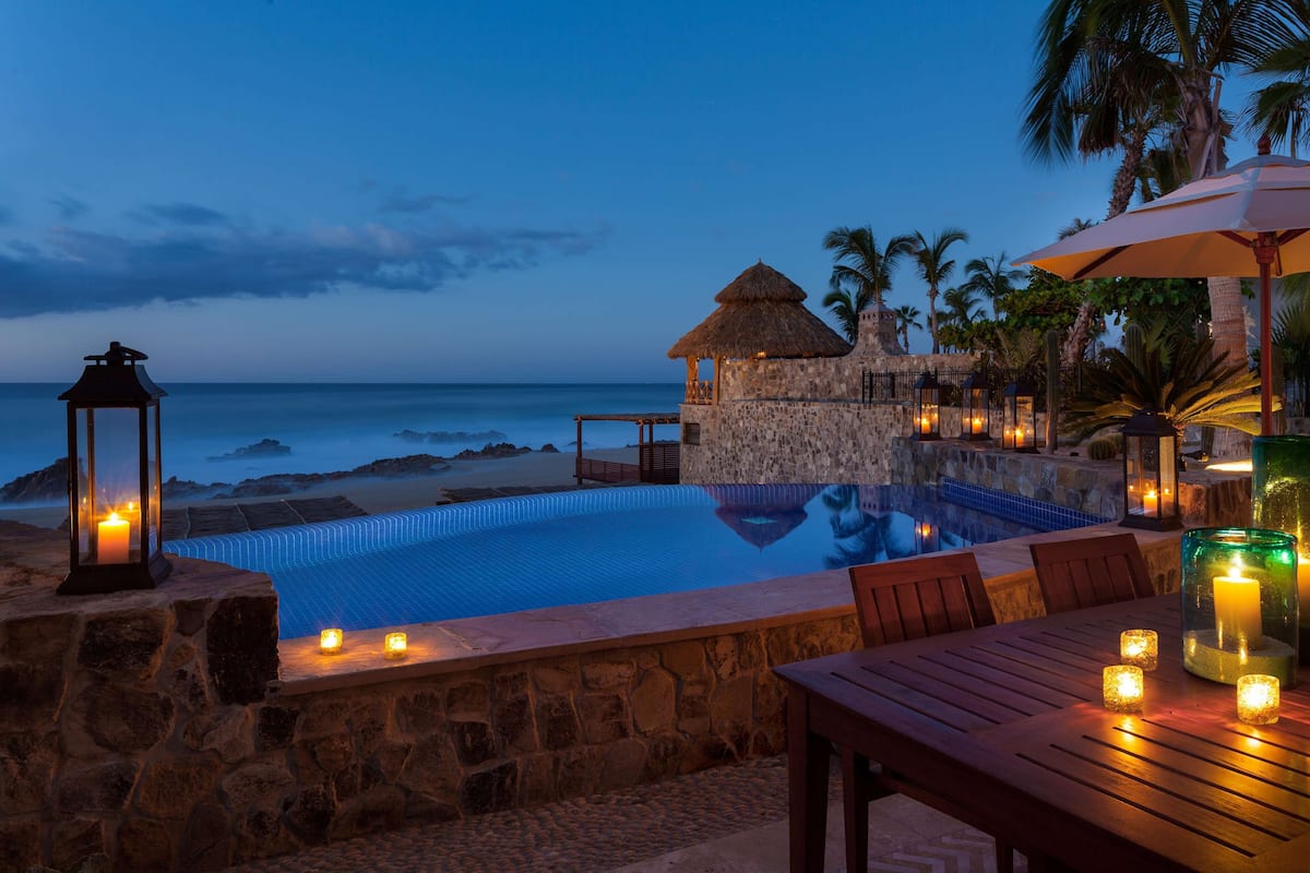 An infinity pool with a stone wall, a thatched-roof structure, and a beach view.