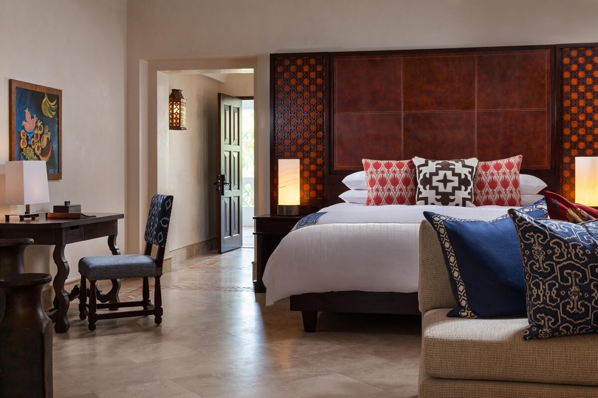 A hotel room with a large bed, a desk, a chair, and a sofa. There is a door leading to a balcony.