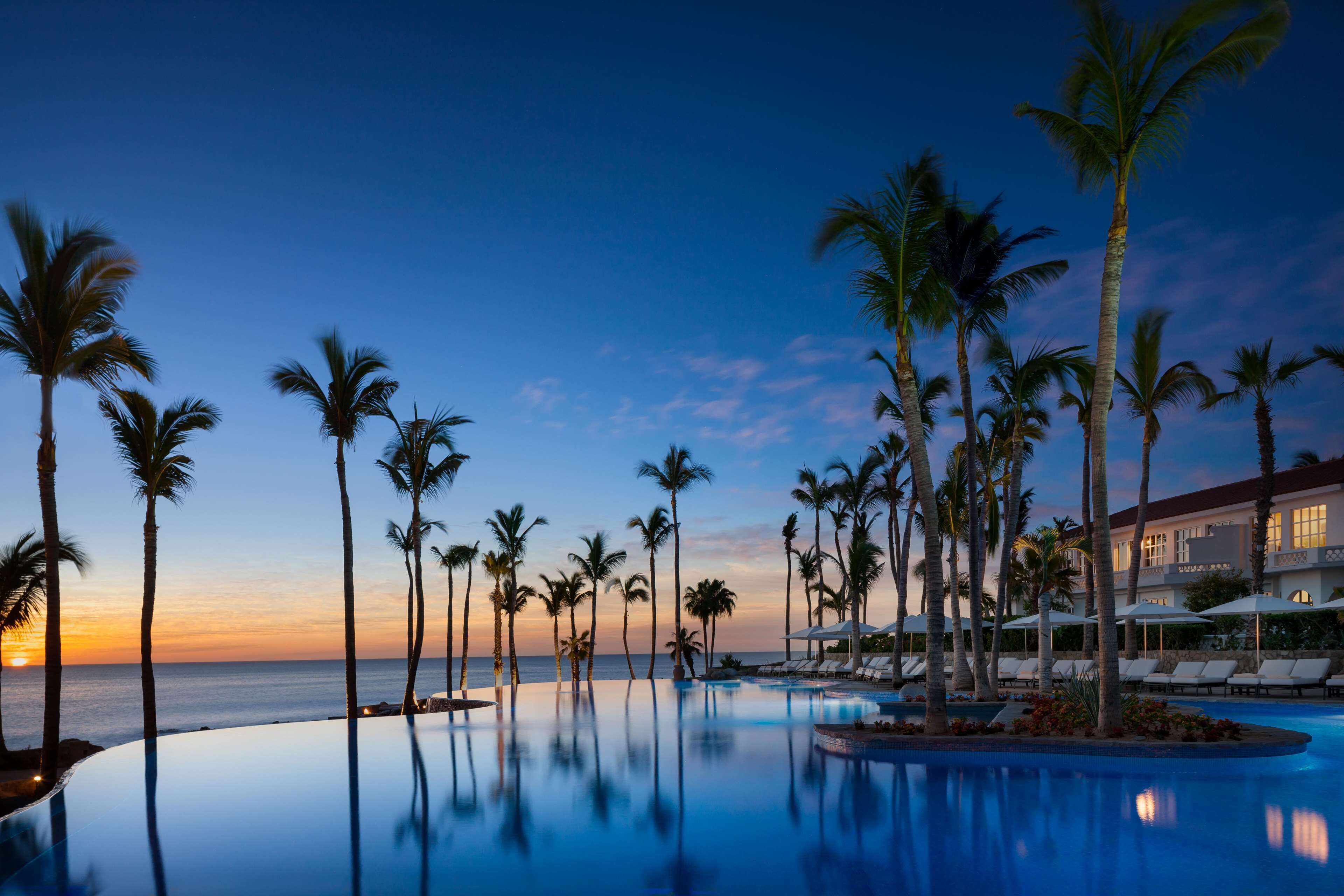 A pool with palm trees and a view of the ocean at sunset.