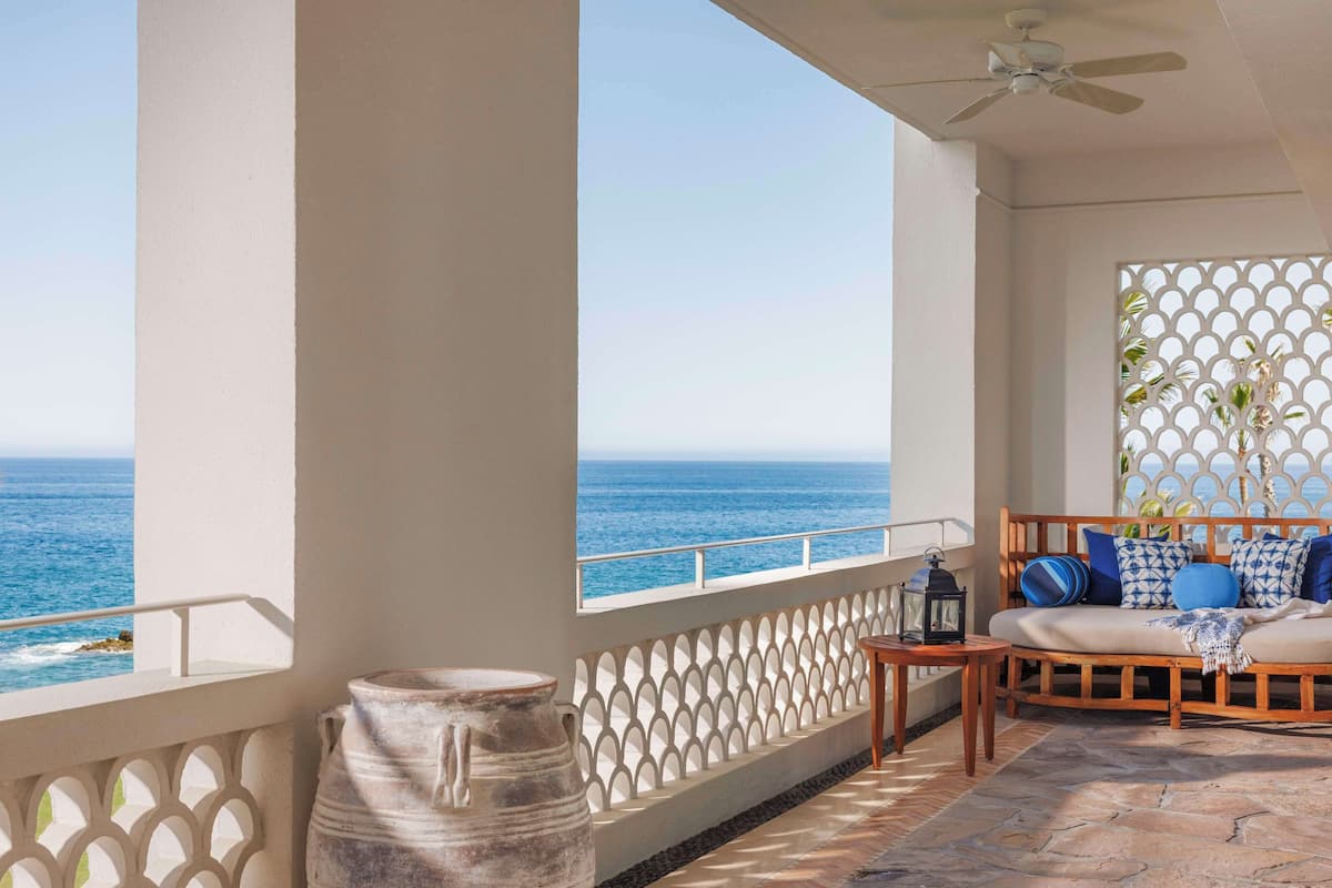 A balcony with a view of the sea, a wooden sofa with cushions, a lantern, and a large vase.