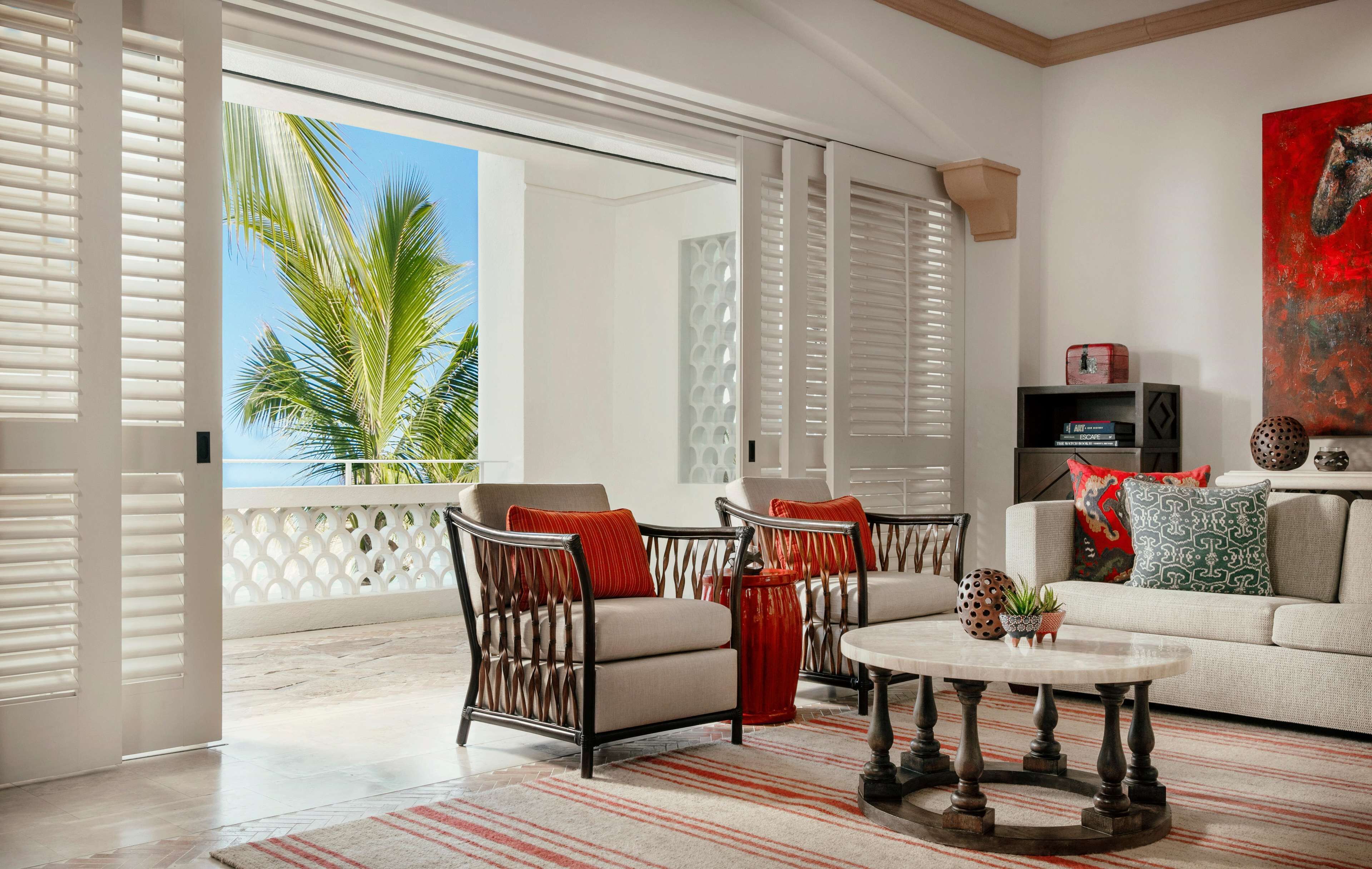 A spacious living room with a sofa, two armchairs, a coffee table, and a balcony with a view of palm trees.
