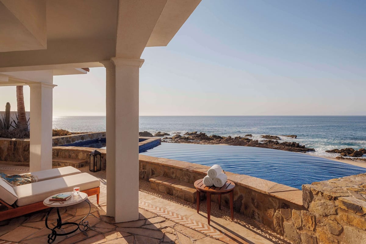 A poolside area with a view of the ocean, a lounge chair, a small table with towels, and a stone wall.