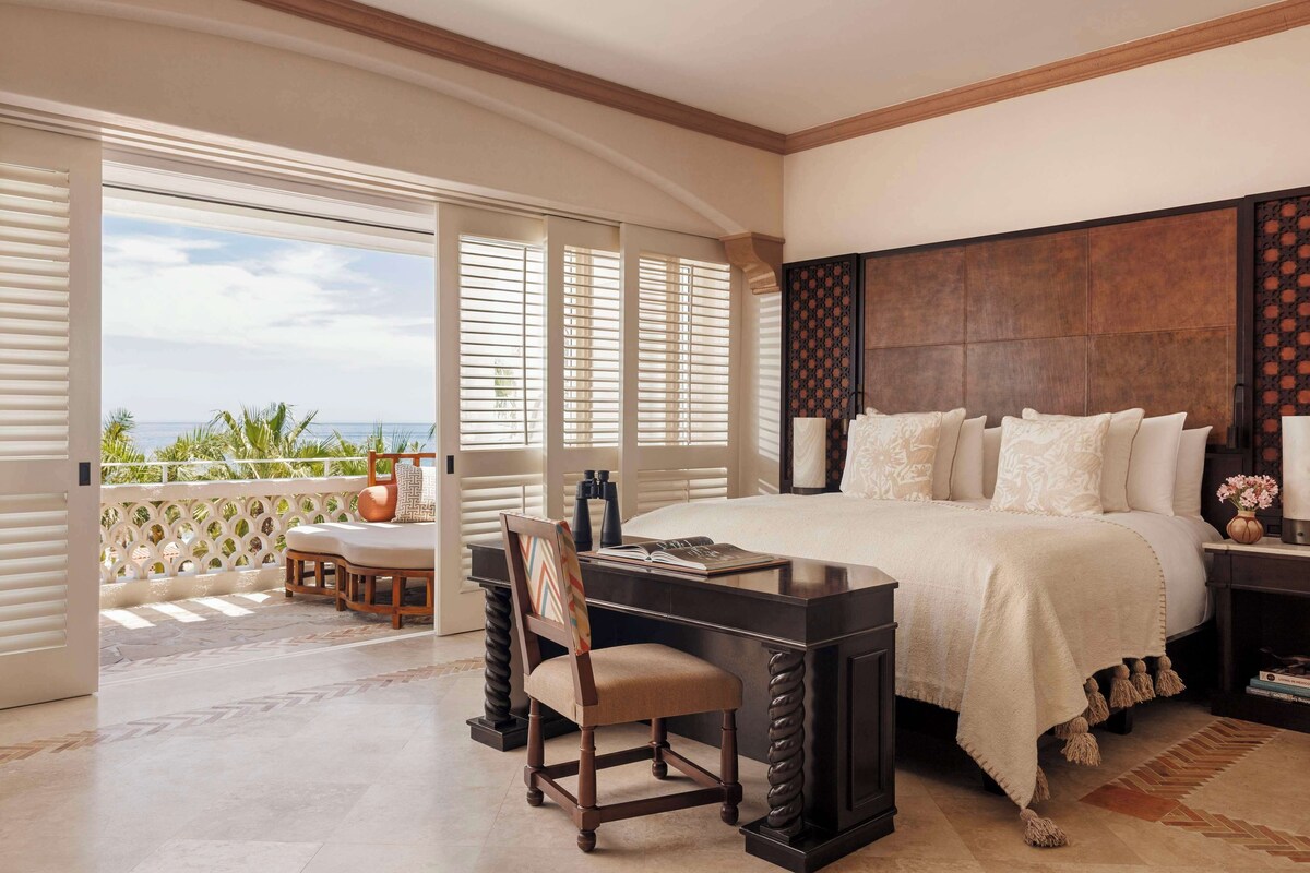 A bedroom with a large bed, a desk, and a chair. There is a balcony with a view of the ocean.
