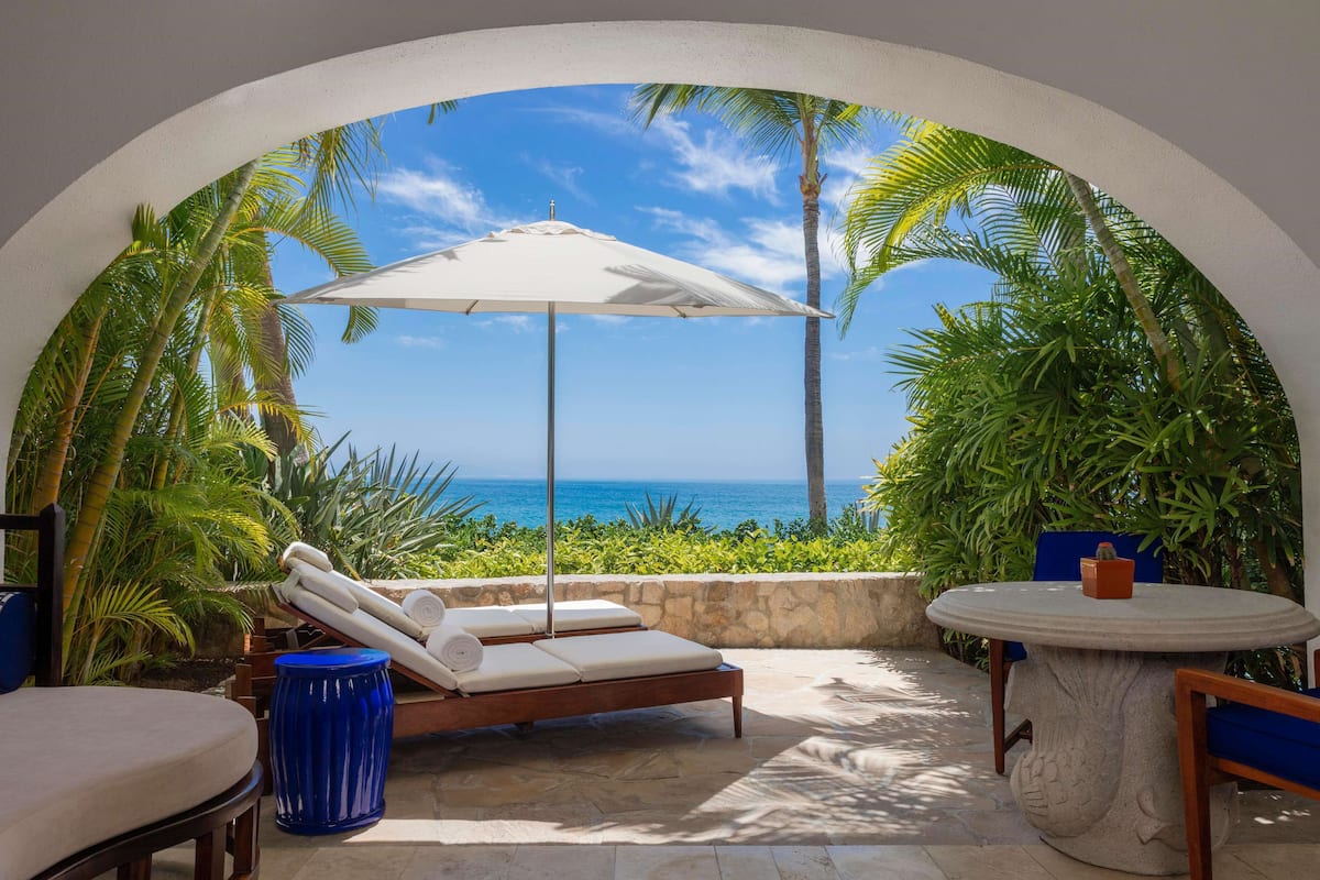 A sunlit patio with a lounge chair, a table, and a view of the ocean through an archway.