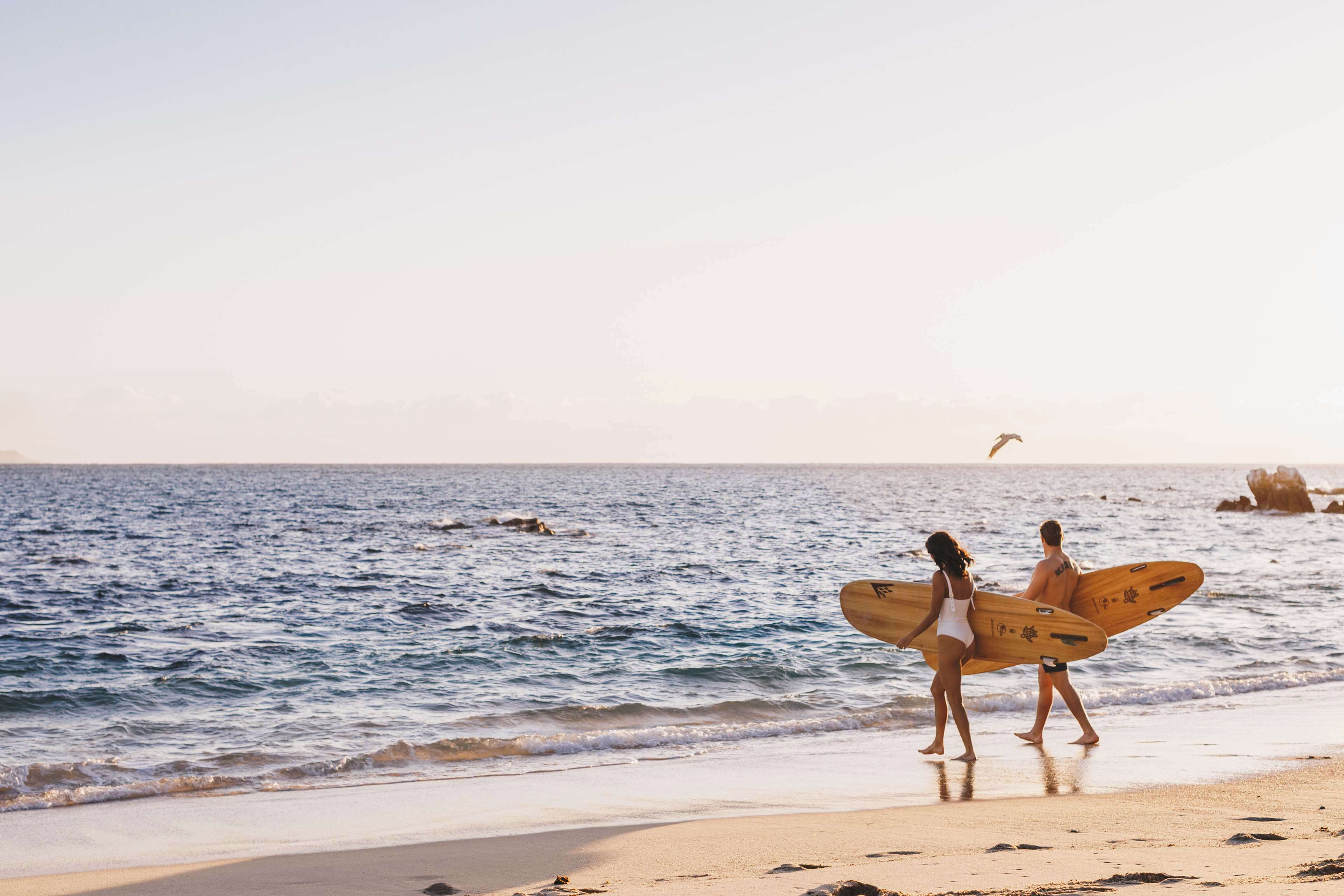 A beach scene with two people carrying surfboards, a clear sky, and calm sea.