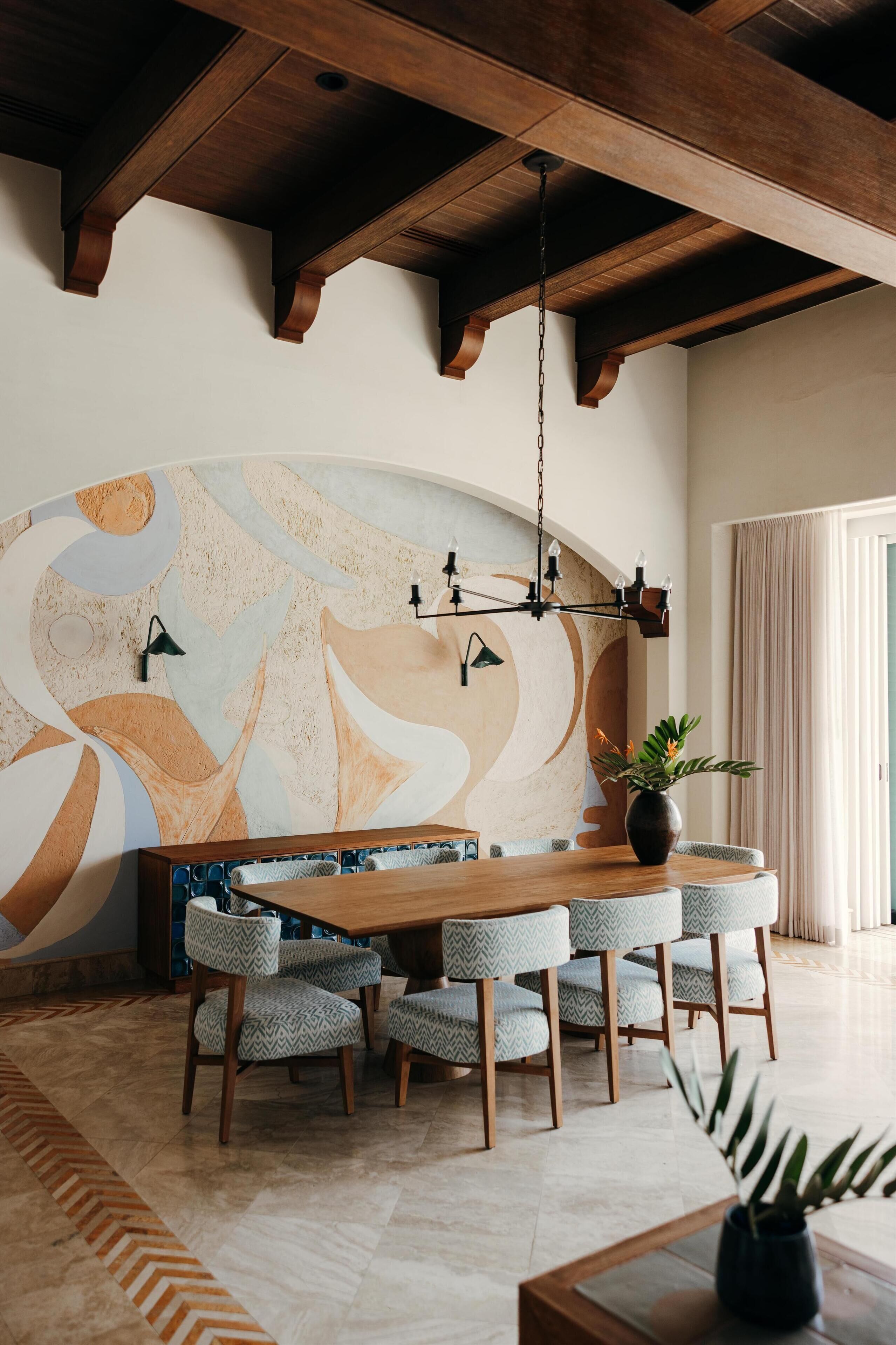 Un espace repas comprenant une table et des chaises en bois, une grande fresque murale abstraite et un luminaire suspendu.
