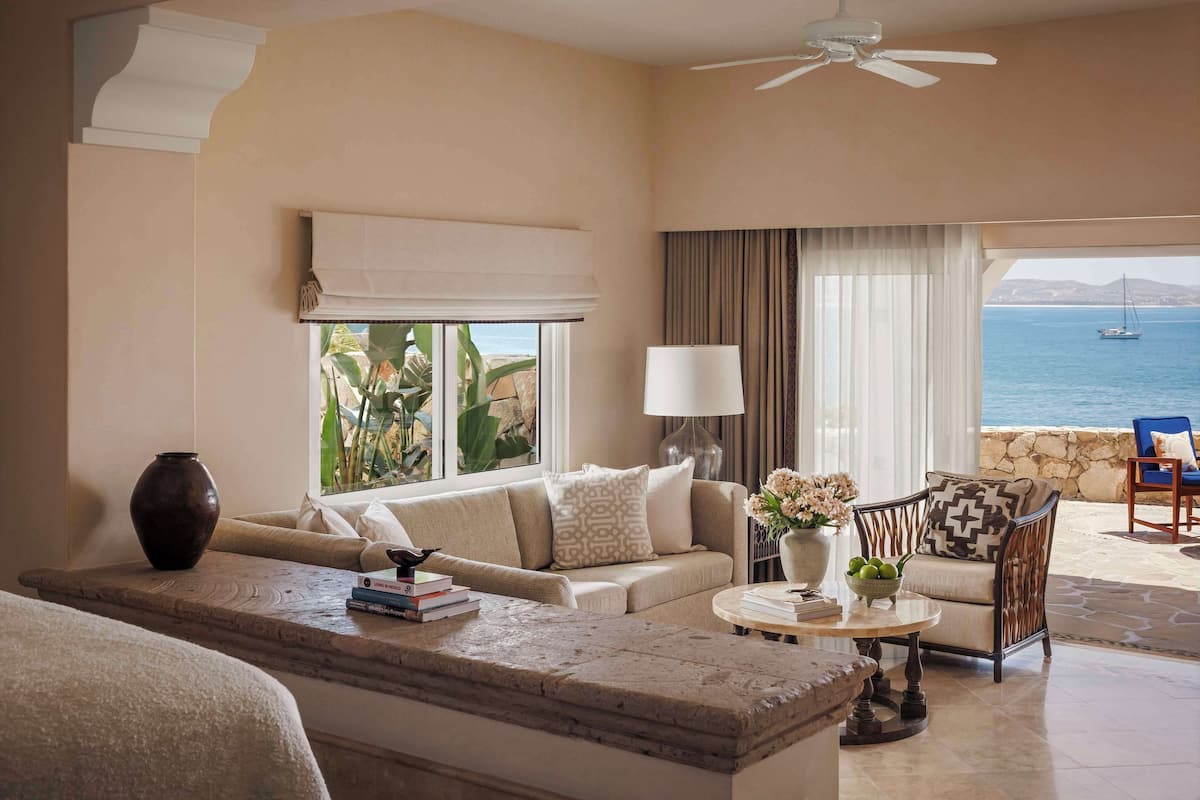 A hotel room with a sofa, armchair, and a dining table. There is a view of the sea through large windows.