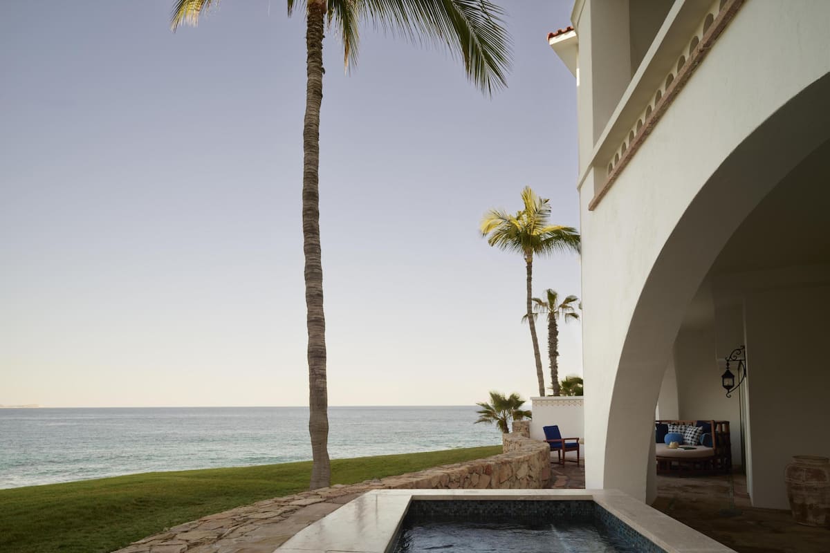 A pool with a view of the ocean, palm trees, and a building with an arched doorway.
