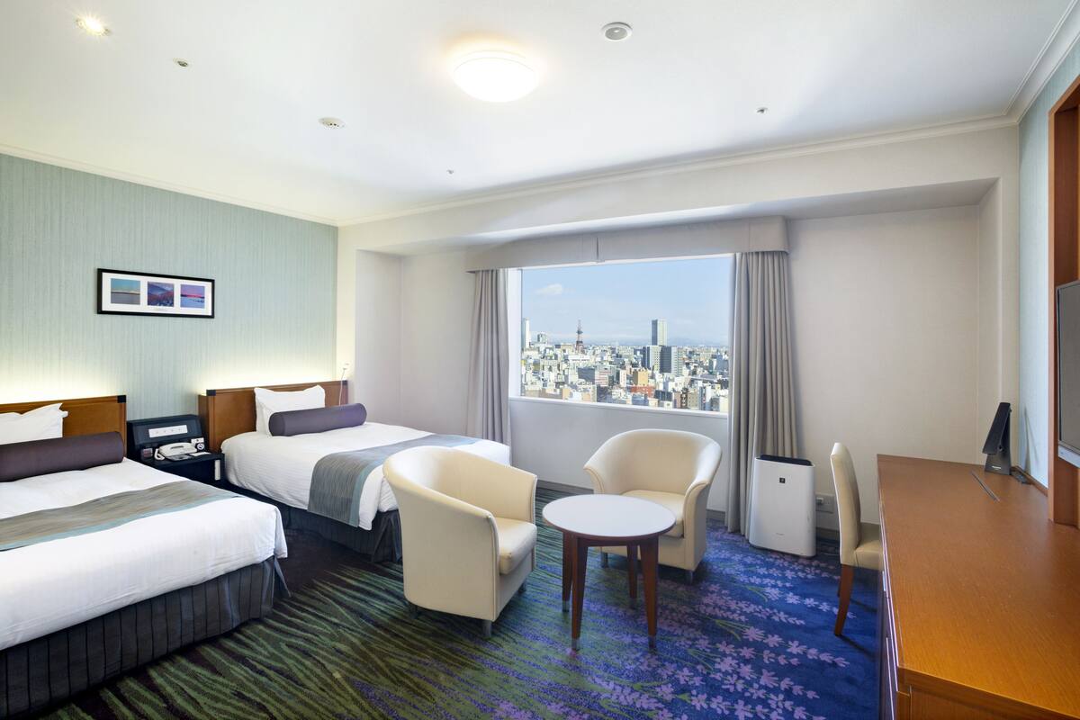 Ein Hotelzimmer mit zwei Betten, einem Schreibtisch, einem Stuhl und Blick auf eine Stadtlandschaft.
