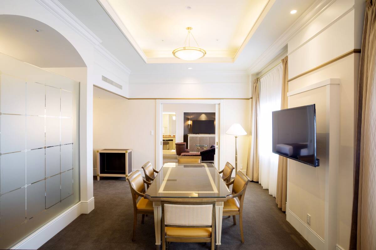 Ein Hotelzimmer mit großem Esstisch, bequemen Stühlen, einem an der Wand montierten Flachbildfernseher und einer gemütlichen Sitzecke mit Sofa und Lampe.