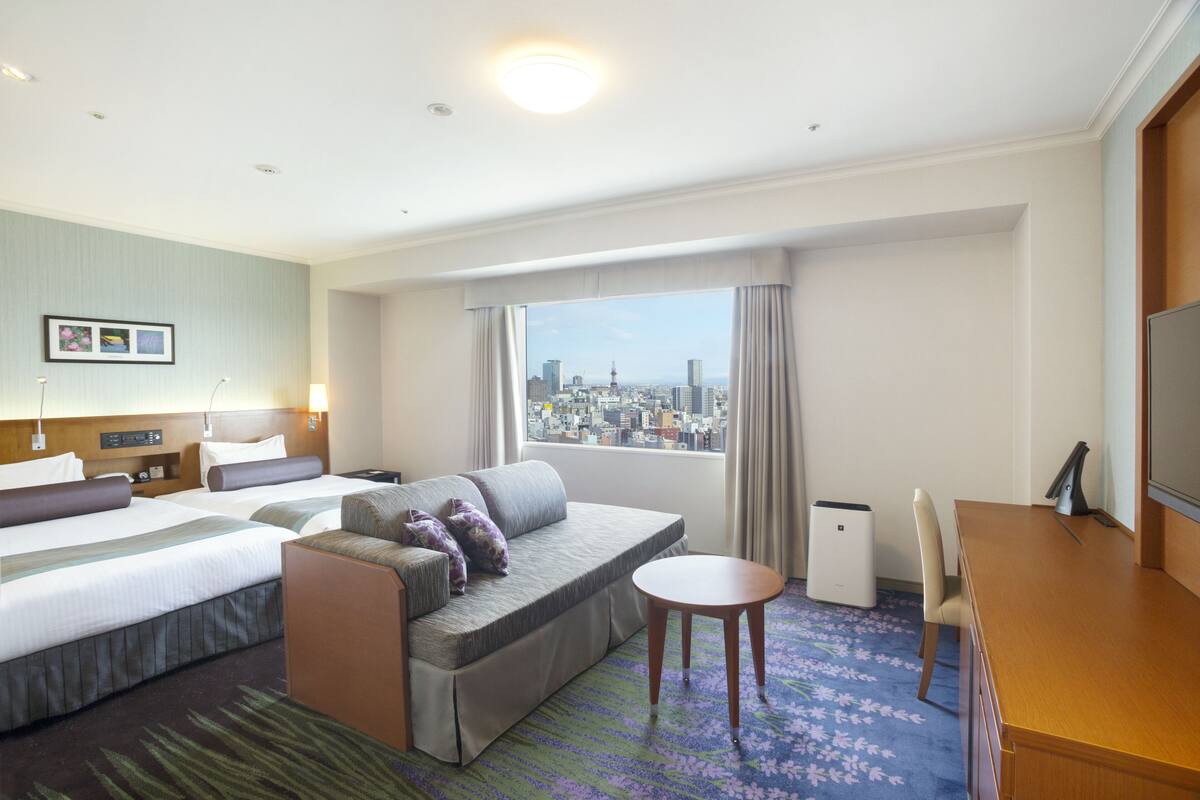 Ein Hotelzimmer mit zwei Betten, einer Sitzecke, einem Schreibtisch und Blick auf die Stadt.