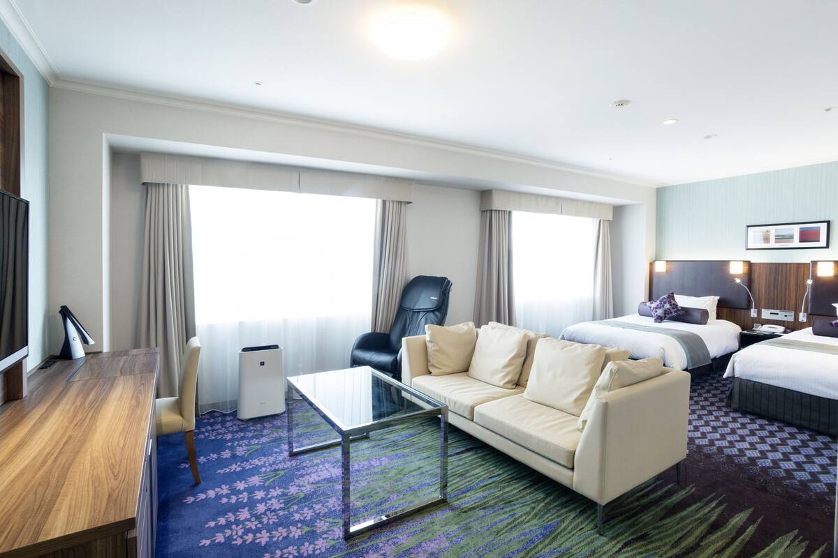 Ein Hotelzimmer mit Schreibtisch aus Holz, einem Glastisch, einer Couch, zwei Betten, einem Sessel und einem großen Fenster mit Vorhängen.