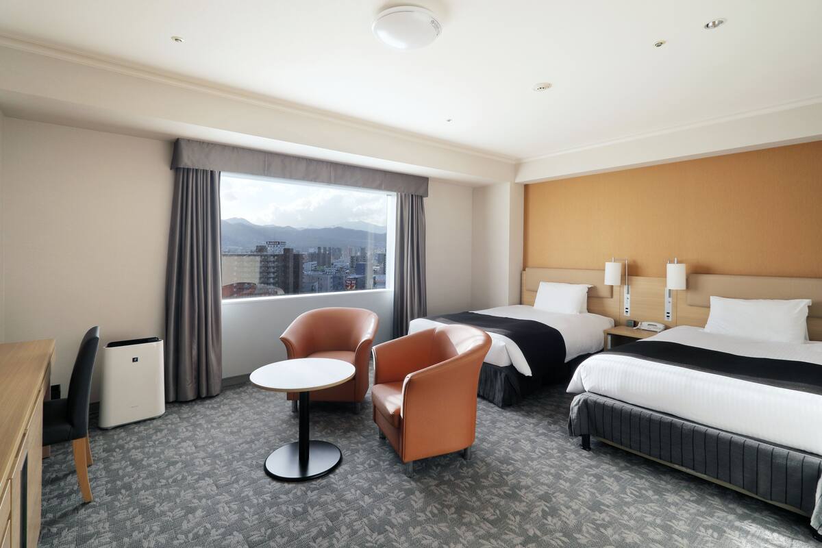 Ein Hotelzimmer mit einem Bett, zwei Stühlen, einem kleinen Tisch, einem Nachttisch und Blick auf eine Stadt durch ein großes Fenster.