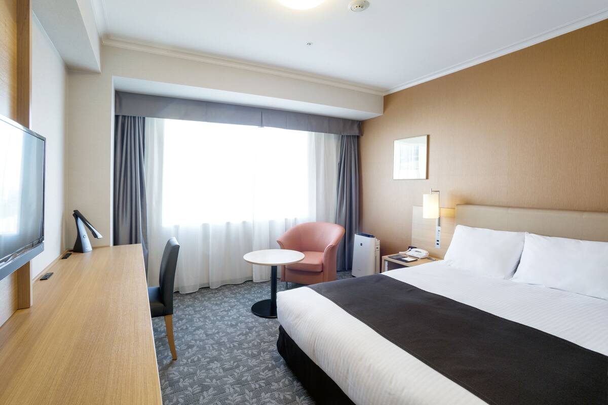 Ein Hotelzimmer mit Bett, Schreibtisch, Stuhl, Fernseher, Lampe und einem Fenster mit Vorhängen.