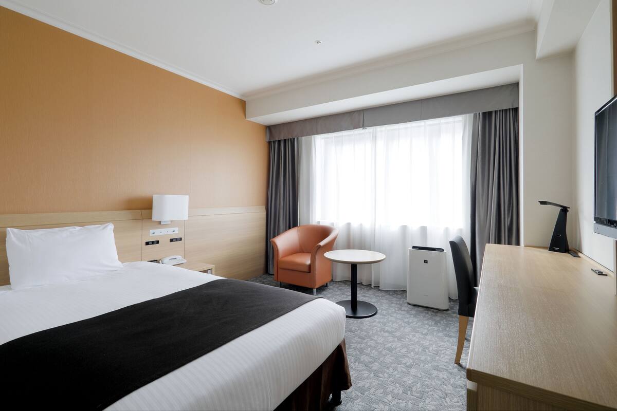 Ein Hotelzimmer mit Bett, Schreibtisch, Stuhl, kleinem Tisch, Lampe, Fernseher und einem Fenster mit Vorhängen.