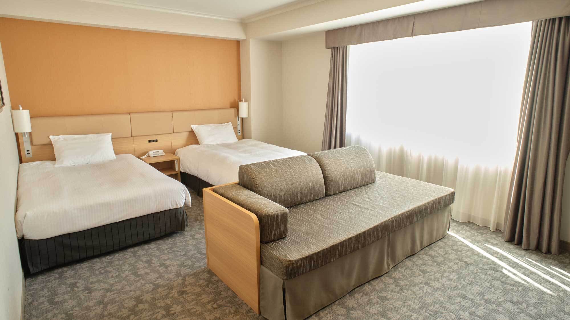 A hotel room with two beds, a sofa, a bedside table with a lamp, and a large window with curtains.
