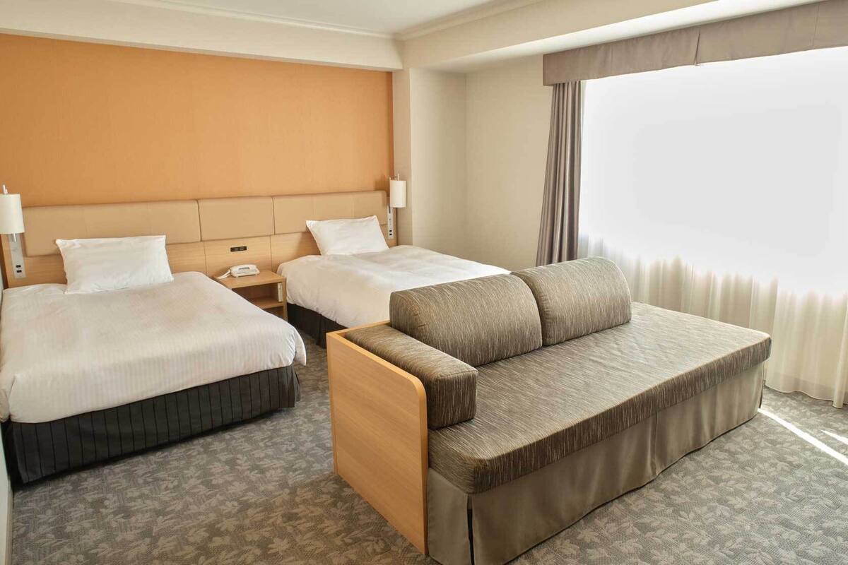 Ein Hotelzimmer mit zwei Betten, einem Schlafsofa, einem Nachttisch mit Lampe und einem großen Fenster mit Vorhängen.