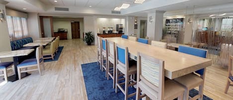 A spacious dining area with a long wooden table and blue cushioned chairs, a separate seating area with a sofa and chairs, and a kitchen area visible through an open door.
