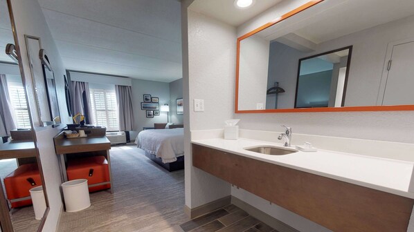 A hotel room with a large bed, a desk with a chair, a vanity with a mirror, and a window with curtains.