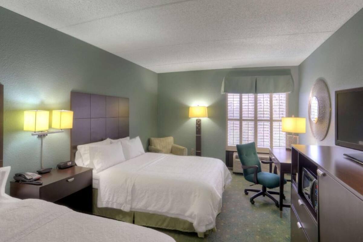 A hotel room with two beds, a desk, a chair, a TV, and a window with blinds.