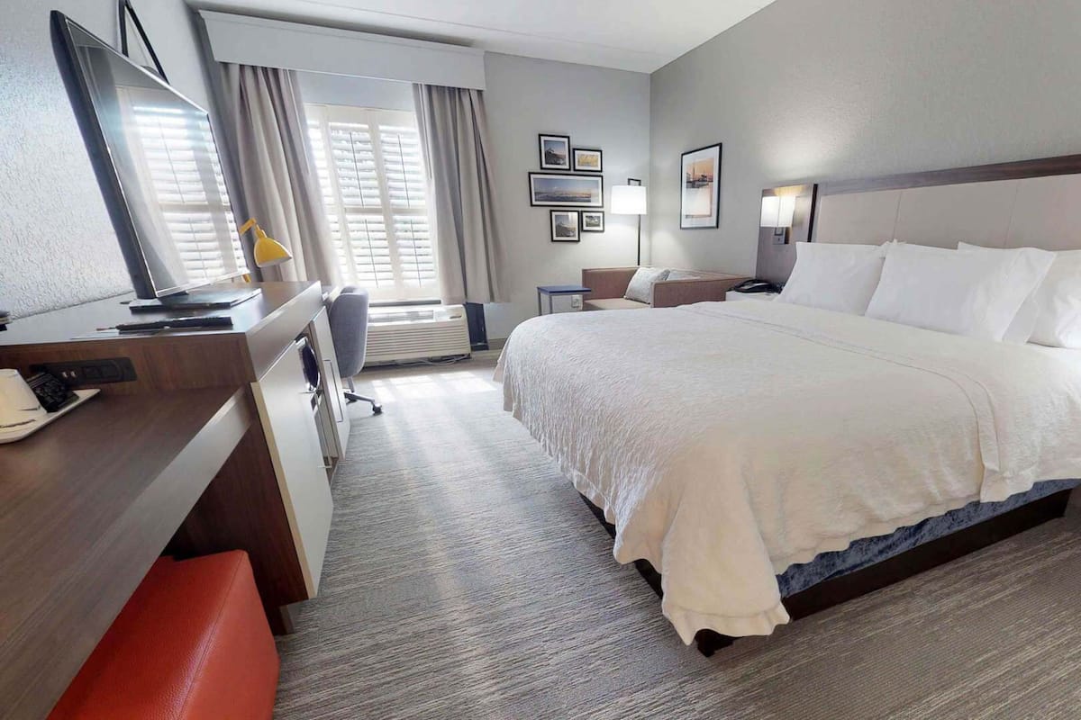 A hotel room with a large bed, a desk with a computer, a chair, a bedside table with a lamp, and a window with curtains.