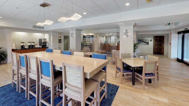 A spacious dining area with a large wooden table and chairs, a coffee station, and a view of the city through large windows. - Hampton Inn St. Simons Island (St. Simons Island)
