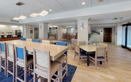 A spacious dining area with a large wooden table and chairs, a coffee station, and a view of the city through large windows.